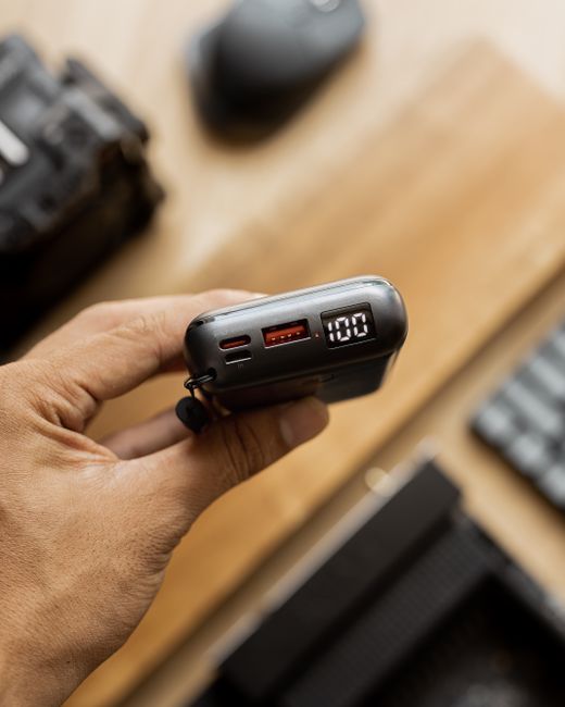 A close-up view of a person's hand holding a portable digital power bank with a digital display showing '100', placed on a wooden surface among various electronic items. Visible around are a camera, a computer mouse, and a keyboard. In the background, a cardboard box and plastic wrapping materials are partly visible, indicating an ongoing packing or packing process. The scene is lit with natural light, suggesting an indoor setting during packing or home relocation activities. This image supports information about packing, moving logistics, and equipment used during house removals, as handled by Man and Van Catford in their furniture transport and home relocation services, aligned with the service category of removals.
