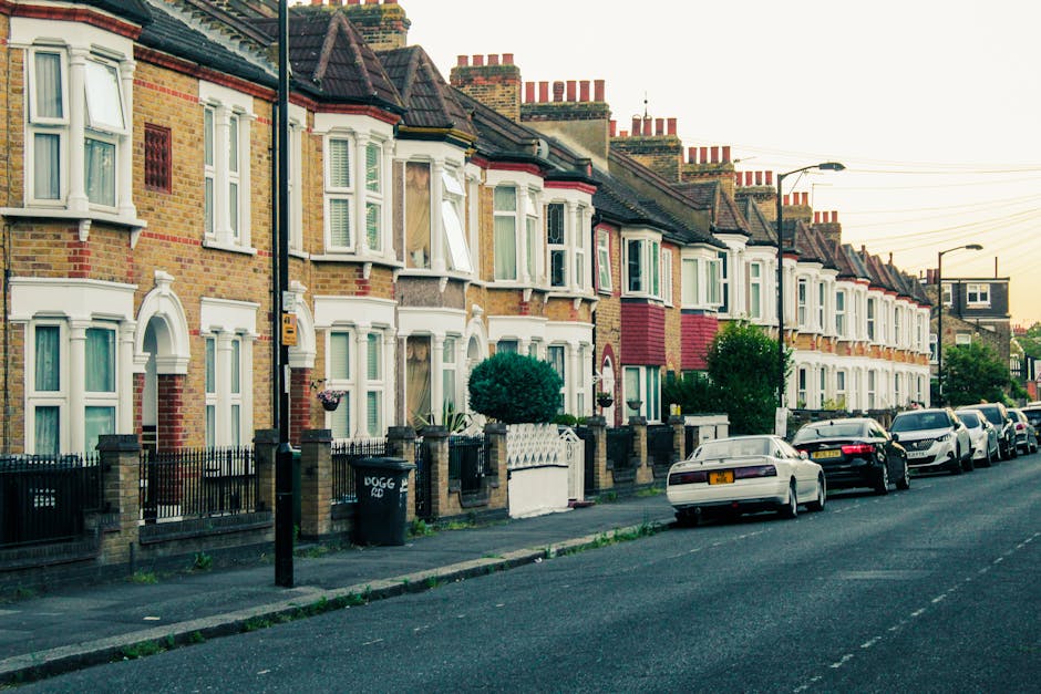 A row of terraced residential houses with brick facades and bay windows on a street in Catford, SE6. The houses have white window frames and decorative elements, with some featuring red brick or painted sections. In front of the homes, there are small front gardens enclosed by low brick walls and iron gates, with neatly trimmed bushes and potted plants visible. Parked along the street are various vehicles, including sedans and hatchbacks, some with visible roof racks. The road is paved and lined with street lamps, with overhead power lines crossing above. Near the sidewalk, a black waste bin is present, and the street appears quiet with no pedestrians visible. This setting provides an urban residential environment, typical for house relocations or furniture transport processes, as conducted by local removal services like Man and Van Catford, ensuring efficient packing, loading, and home relocation activities.