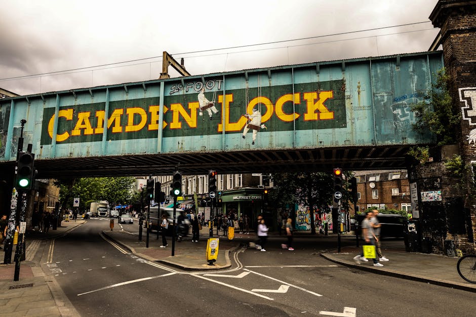 A large, weathered railway bridge spans over a busy street intersection in Camden, with the words '20 FOOT' painted above the word 'CAMDEN LOCK' on the side of the bridge in bold yellow letters. The b