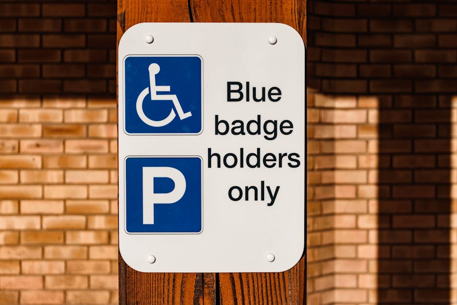 A rectangular white parking sign mounted on a wooden post, with two blue symbols and black text. The top blue symbol displays a white wheelchair icon, indicating accessible parking, and below it, the