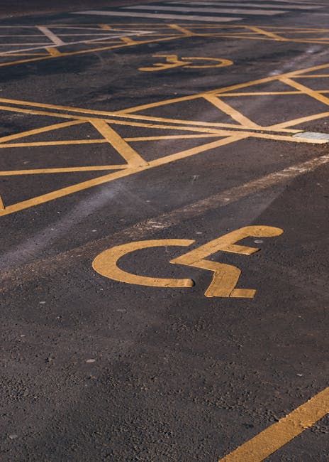 A rectangular white parking sign mounted on a wooden post, with two blue symbols and black text. The top blue symbol displays a white wheelchair icon, indicating accessible parking, and below it, the